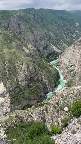 The video displays a mountainous canyon from a high vantage point. The canyon walls are steep and rocky, with green vegetation covering parts of the slopes. A river flows through the bottom of the canyon, its waters appearing turquoise in color. The sky above is partly cloudy, with patches of blue visible. There are no people or animals visible in the frame. The camera is mostly still, providing a wide shot of the landscape.