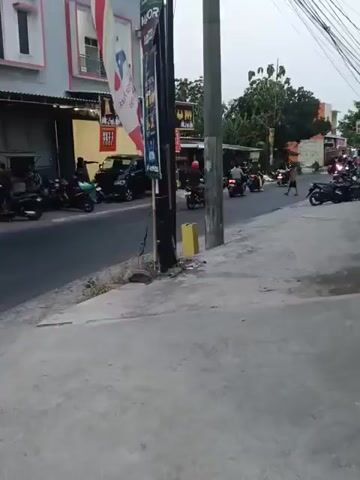 The video shows a street scene with several motorcycles parked along the side of the road. A few people are visible, some near the motorcycles and others walking. There are buildings lining the street, with storefronts and awnings. Power lines are visible overhead. The time of day appears to be late afternoon or early evening, based on the lighting. The overall atmosphere is that of a busy street in a commercial area.