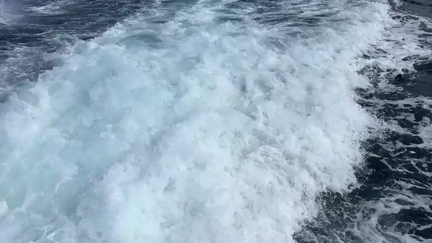The video shows a wake of white water trailing behind a moving boat. The water is dark blue and choppy. In the distance, a coastline with hills and buildings can be seen under a partly cloudy sky. The camera appears to be positioned low on the boat, looking back at the wake and the receding shoreline.
