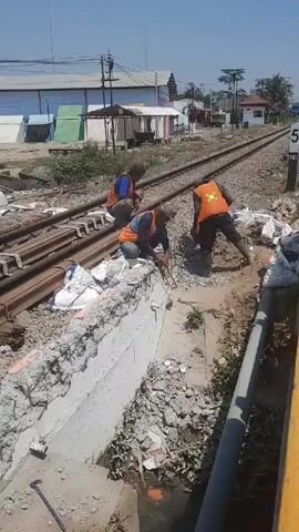 Three individuals wearing orange safety vests are working on a construction site near a railway track. The ground is covered with gravel and debris. In the foreground, a concrete structure is being worked on, with rebar visible. One individual is using a tool, possibly wire cutters, to work on the rebar. Another individual is kneeling beside them, and the third individual is standing further away, appearing to be working with a tool as well. In the background, there are railway tracks, some buildings, and vegetation. The sky is clear and the scene is well-lit, suggesting it is daytime.