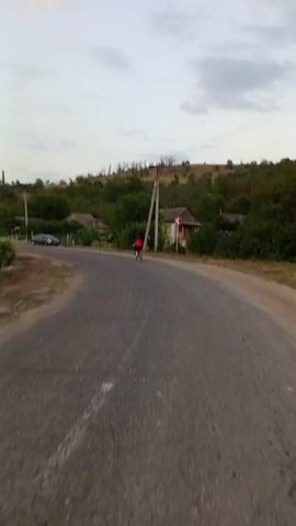 A person is riding a bicycle on a paved road through a rural area. The road curves to the left and goes uphill. There are houses and trees on either side of the road, and telephone poles are visible in the distance. The sky is cloudy and the light suggests it is either early morning or late evening. The person on the bicycle is wearing a red shirt and is in the middle of the road. A car is parked on the side of the road to the left. The scene is observed from a first-person perspective, as if the viewer is also on the road, possibly running or walking.