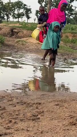 Three people are wading through shallow water in a rural, outdoor setting. A woman wearing a bright pink headscarf carries a small child on her back. Another child, wearing a green cloak, walks in front of her, carrying a yellow bucket. The water reflects the sky and trees. The ground is mostly dirt and sand, with some grass visible in the background. The sky is overcast.
