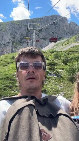 A man and a girl are riding a ski lift up a mountain. The man is in the foreground, wearing sunglasses and a white t-shirt. He is holding a brown bag. The girl is to his right, partially visible. The ski lift is moving upwards, with chairs visible above them. The mountain is covered in green grass and rocky cliffs. The sky is blue with some clouds. The video appears to be a selfie taken by the man.