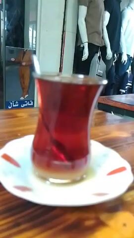 Certainly! Here's a description of the video:

The video focuses on a traditional Turkish tea glass filled with dark red tea and a small spoon, sitting on a white saucer with red teardrop designs. The cup and saucer are placed on a wooden table with a visible grain pattern. The background is somewhat blurred, but appears to show the interior of a clothing store, with mannequins dressed in various outfits. A sign with Arabic script is visible in the background as well. The camera is static, offering a close-up shot of the tea.