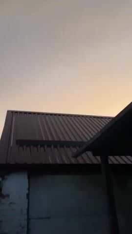A metal roof is shown against a pale sky during what appears to be dusk or dawn. The roof is dark and corrugated, with the sun casting a warm glow on the sky. Part of a building is visible below the roof, and it appears to be a residential or industrial structure. There are no people or animals visible in the video.