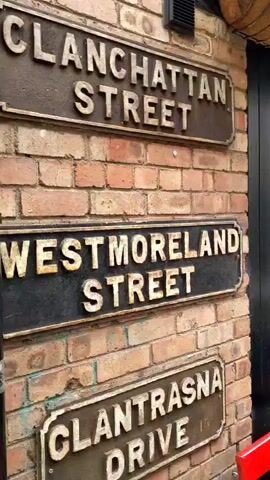 The video opens with a close-up of three weathered metal street signs attached to a brick wall. The signs read "MANHATTAN STREET", "WESTMORELAND STREET", and "GLANTRASNNA DRIVE". The camera then pans to the right, revealing a narrow cobblestone alleyway lined with buildings. Colorful umbrellas are suspended overhead, creating a vibrant canopy.

On the left side of the alley, a person wearing a white baseball cap and a grey hoodie sits on a red bench, looking at their phone. They are wearing dark pants and have a colorful scarf draped over their lap. A glass of beer is on the bench next to them.

Further down the alley, several other people are visible. Some are sitting on benches outside establishments, while others are walking. The buildings have dark wooden doors and windows, and many are adorned with hanging flower baskets. Signs for "LADIES ROOM", "WHISKY", and "OLD BUSHMILLS WHISKEY" are also visible on the buildings.

The overall atmosphere appears to be casual and social, with people enjoying the outdoor space. The alley is clean and well-maintained, with the cobblestone pavement and brickwork suggesting an older, historic area. The lighting suggests it is daytime, though the umbrellas cast shadows, creating a dappled effect.