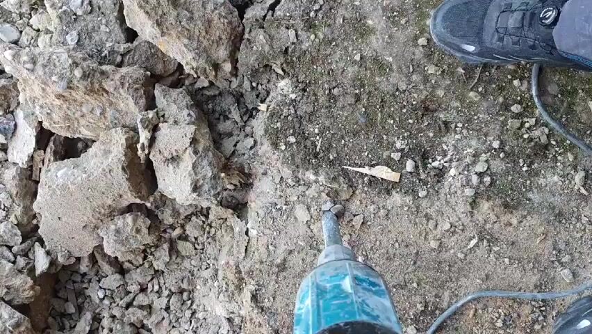 The video shows a teal-colored jackhammer breaking apart a pile of grey, gravelly rock. The jackhammer is being operated by a person, visible by their black sneakers in the upper right corner of the frame. The camera angle is from above, looking down on the tool and the surface being worked on. The area surrounding the rock pile consists of dirt and small gravel.  A power cord extends from the jackhammer.