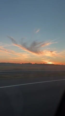 A car drives along a road with a scenic sunset in the background. The sky is a gradient of blue and orange, with wispy clouds catching the last rays of the sun. The road is asphalt with white lines marking the lanes. The car is white and appears to be a sedan. The overall scene is peaceful and serene.