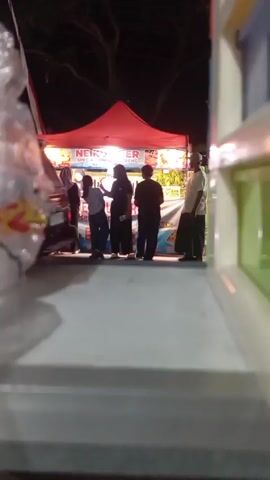 The video shows a nighttime scene at an outdoor food stall with a red canopy. Several people are visible in front of the stall, some appearing to be customers and others potentially working there. The camera angle is low, as if looking out from behind a counter or table. The lighting is dim, with the stall's lights providing the primary illumination. Some items are visible on the counter in the foreground, including what appears to be a clear plastic bag on the left. The background is dark with some trees visible above the canopy. The people are mostly silhouetted against the stall's lights, making detailed identification difficult. One person in the center appears to be wearing a dark headscarf and a dark dress. Another person on the far right is wearing a light-colored shirt and pants and a cap. There is some movement among the people as they interact. The overall impression is of a casual, busy evening at a local food vendor.