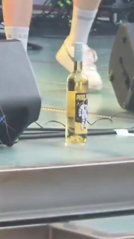 The video shows a live performance on an outdoor stage. In the foreground, a bottle of white wine stands on the stage floor. Behind it, a man with a beard and a dark t-shirt and shorts is singing into a microphone, with one leg resting on the bottle. To his right, another person in a dark t-shirt and shorts, wearing a baseball cap, is bending down and pointing. To the left, a drummer is visible behind a drum set. The stage is made of wood and metal, with speakers and lighting equipment set up. The background includes a white tent, bamboo decorations, and a sign that partially reads "Ho Sum". The overall scene is casual and appears to be a summer event.