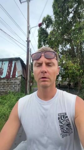 The video features a close-up, chest-up shot of a fair-skinned man, likely in his 30s or 40s, with short, light-colored hair. He is wearing a plain white sleeveless shirt or tank top, which has "ULUWATU" printed in black text on the lower right side. A pair of sunglasses with reddish-brown frames is pushed up onto his forehead, reflecting the cloudy sky.

The man looks directly at the camera throughout the video. His expression is initially serious and somewhat tense, with his mouth slightly agape. As the video progresses, a subtle, almost imperceptible smile begins to form on his face.

The background indicates that the man is outdoors and appears to be on a moving vehicle, possibly a scooter or motorcycle, as the scenery shifts rapidly. The setting is a rural or semi-rural area under a cloudy sky. Dominant background elements include:
- Numerous overhead power lines and a utility pole stretching across the frame.
- On the left side, an old, rustic shack or small building with a corrugated metal roof and brick walls is visible in the initial seconds.
- Beyond the immediate foreground, there are green fields, possibly rice paddies or overgrown grass.
- Various houses and structures, some with traditional Southeast Asian architectural styles (like a gate with intricate carvings), appear in the distance on both sides of the road.
- A white sign with some dark, indistinct text is briefly visible on the right side of the frame.
- Scattered trees and lush green foliage complete the outdoor tropical environment.

The man speaks several words, which sound like "Воронок" (Voronok) and "Карман" (Karman) in Russian, repeating the latter phrase multiple times. The video is filmed during the daytime, under an overcast sky, providing soft, diffused lighting.