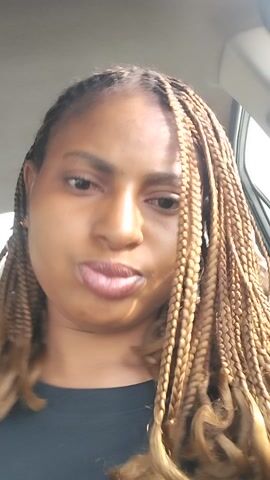 A young woman with brown braided hair and glossy lips sits in a car. She is wearing a black shirt. The sun shines on her face, creating a light and shadow effect. She is holding her phone to record the video. The interior of the car is visible, including the door and seat.