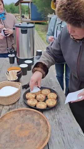 The video shows a group of people gathered outdoors at a wooden table. A woman wearing a fur hat and brown coat is picking up small pies from a black plate with a white napkin and handing them out to the people standing around the table. There is a large stainless steel coffee urn, paper cups, wooden stirring sticks, and a small basket containing white sugar on the table.  One person wearing a pink puffer jacket and jeans is talking on a cell phone and holding a paper cup. Another person wearing a grey and white jacket is holding a paper cup with a beverage in it, and taking a pie from the woman in the fur hat. In the background is a wooden pavilion, trees, and an open grassy area.
