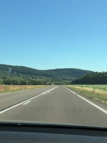 This is a first-person perspective video clip of driving on a paved road through a rural, hilly landscape under a clear blue sky. The road has a white dashed line on the right edge and solid white lines marking lanes. A white arrow indicating a left turn is painted on the road surface in the foreground.

The scene is characterized by lush green hills covered in dense trees, with a field of tall, dry grass or grain along the right side of the road. In the distance, more hills stretch across the horizon, and a few distant vehicles are visible on the road ahead. The sun is high in the sky, suggesting it is daytime, likely mid-morning or afternoon, and the weather appears to be warm and clear, typical of summer in France.

There are no people or distinct objects other than the road and the natural landscape. The setting is serene and suggests a leisurely drive through the countryside. The Villages du Lac de Paladru, France is mentioned as the location context, implying the drive is in this region. The overall mood is peaceful and calm, with a sense of journey and exploration. No text is visible in the video.