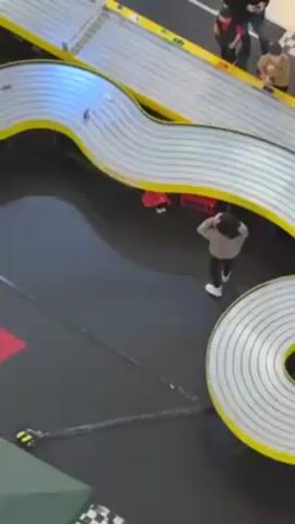 A high-angle shot shows a large slot car race track in a mall or similar venue. The track, outlined in yellow, forms a complex, winding path. A person stands near the track, adjusting or observing a red slot car. Onlookers are present on the far side of the track. The black surface around the track features "MONZA" in red letters. Green umbrellas and a checkered pattern decorate the area.