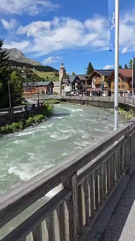 A video shows a scenic view of a town situated alongside a fast-flowing river. Traditional wooden chalets and buildings with pointed roofs line the riverbanks, with a church steeple visible in the background. Mountains rise behind the town, covered in green vegetation and with a few patches of snow on the higher slopes. The sky is blue with fluffy white clouds. The video is shot from a wooden bridge or railing, looking out over the river towards the town. A flagpole stands to the right of the frame, with a flag partially visible at the top. The water in the river is a milky, turquoise color, and appears turbulent with small waves and rapids. The overall impression is of a picturesque mountain village on a sunny day.