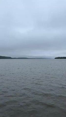 The video shows a wide expanse of choppy gray water under an overcast sky. In the distance, a low, tree-covered shoreline is visible on the left, and another stretch of land is visible on the right. Farther in the distance, across the water and along the horizon, there are some faint, white structures that could be buildings or ships. The camera appears to be on a boat moving slowly through the water. The overall atmosphere is somber and muted.