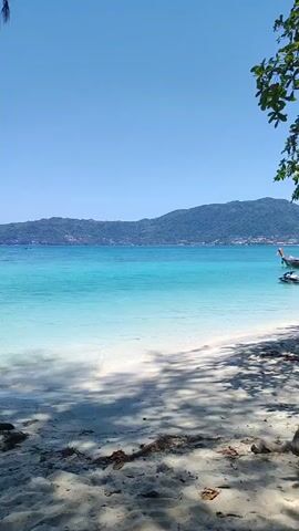 The video shows a bright, sunny day at a tropical beach. The sand is white, and the water transitions from clear near the shore to a vibrant turquoise further out. Across the water, there is a hilly island or landmass. Some vegetation frames the scene on the sides and at the top, casting shadows on the sand in the foreground. There's minimal activity, emphasizing the tranquility of the location.