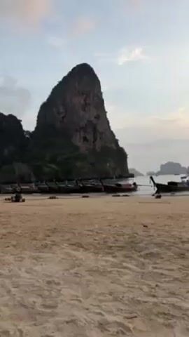 The camera pans across a sandy beach with a large rock formation covered in greenery on the left. Several longtail boats are docked near the rock. A person walks along the beach towards the ocean where another person is swimming. The sun is setting, casting a warm glow on the water and sky, which is partially covered by clouds.