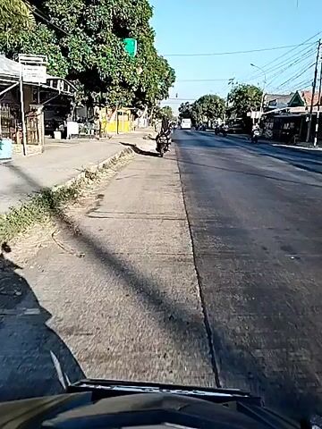A first-person perspective video shows a motorcycle traveling down a paved road. The road is lined with trees and buildings on both sides. The sky is clear and blue. Several other motorcycles and a van are also visible on the road, traveling in the same direction. The motorcycle is moving at a moderate speed, and the driver appears to be alone. The video is recorded in daylight, and the lighting is bright. The overall atmosphere of the video is calm and peaceful.