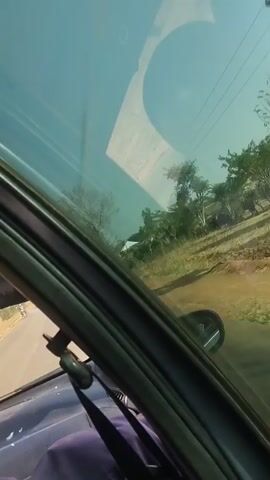 The video shows a view from inside a moving car. The car is traveling on a dirt road through a rural area. There are trees, dry grass, and a few small buildings visible outside the car window. A power line with poles is also visible. The car's side mirror is visible in the window's reflection. Inside the car, the side of the seat, a seatbelt, and part of the car's interior trim are visible. The sky is light blue and visible through the car window. The video is slightly shaky, indicating it was likely filmed by hand.
