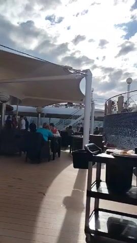 The video shows an outdoor restaurant area on a cruise ship. The camera focuses on a two-tiered service cart with food and drinks on the top level and black bins on the bottom level. A waiter wearing a white shirt, black pants, a green apron, and a green hat pulls the cart through the restaurant. The restaurant has tables and chairs covered by white awnings. People are seated at the tables eating. In the background, there is a view of the sky with clouds. There is also a railing and a mosaic-tiled wall. The floor is made of wooden planks.