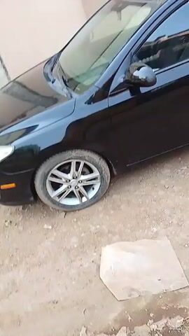 The video shows two black cars parked on a dirt lot, close to a beige wall. The first car is a hatchback. The second car is a sedan, specifically a Toyota Corolla. The sedan has a license plate that reads: BDU-123AA. The video is shot at eye level and the camera moves from the front to the back of the two vehicles. There are no people visible in the video.