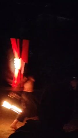 A man is performing with fire in a dark outdoor setting. He is wearing loose dark pants and is shirtless. He is holding a flaming object in his right hand and appears to be swinging it. In the background, red curtains are visible, illuminated by a light that creates a green glow. The ground is dirt or sand. The video is low light and appears to be handheld. There is an audience member in the foreground, silhouetted.