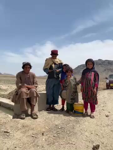 The video captures five individuals, likely a family, in a rural, arid landscape in Ashoqah, Afghanistan, during bright daylight. An older man, dressed in a light brown traditional shalwar kameez and cap, is seated on a low concrete structure to the left. To his right, a taller, younger man or teenager stands, wearing a dark blue traditional outfit, a light shawl, and a red cap, gesturing with his hands.

Beside him are three children. A young girl in a blue headscarf and patterned top holds a metal pot. Next to her, another young child, wearing a green patterned outfit and a light blue scarf, begins to perform a playful dance, swaying and leaning. On the far right, a young girl in a vibrant pink patterned dress and a dark headscarf stands smiling at the camera.

The ground is dry dirt, suggesting a rural road or open area. In the background to the left, dry, possibly fallow, fields stretch towards distant hills or mountains. More rugged mountains are visible in the far background to the right. A vehicle, possibly an SUV, is partially visible on the dirt road in the right background. A yellow bucket with the text "SHARJAH CITY" is visible near the children's feet. The sky is bright with some scattered clouds. The overall atmosphere appears cheerful, with the children engaging playfully.