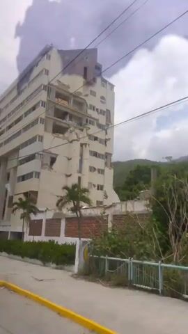 A video pans across the exterior of a damaged, multi-story building. The building's facade shows significant structural damage, with sections of the exterior walls collapsed and broken, revealing the interior floors and rooms. There are visible cracks and deformations in the concrete structure. Palm trees and other greenery are present in front of the building, along with a low brick wall. A yellow car is parked near the building entrance. Power lines are visible in the foreground, and the sky is overcast. The video appears to be captured from a moving vehicle.