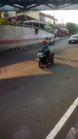 A person rides a black motorcycle down a paved road. The rider wears a blue shirt, dark pants, and a pink helmet. Sunlight casts long shadows across the road. Buildings and a concrete barrier line the right side of the road, and a bridge structure is visible overhead. Another vehicle, a black SUV, travels further down the road, and a white pickup truck is parked on the right side of the road. The scene appears to be in an urban or suburban area during daylight.