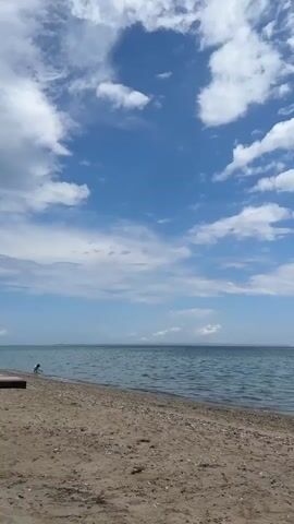 The video transitions from a view of a sunny beach with a blue sky and scattered white clouds to an overcast beach with gray clouds. In the first scene, the ocean is calm. In the second scene, several people are at the beach, some standing in the water and some sitting on the beach.