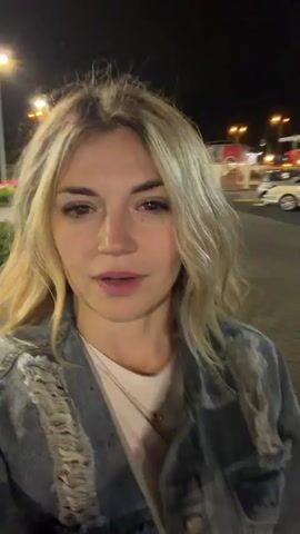 A woman with blonde hair is standing outside of a McDonald's restaurant at night. She is wearing a white t-shirt and a blue denim jacket. She is smiling at the camera and talking, likely recording a video. Behind her is the McDonald's building, with a golden arches logo and "McDonald's" in white letters. There are cars parked in the parking lot in front of the building.
