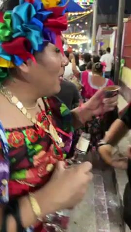 The video shows a person in traditional Mexican dress holding a bottle of liquor as another person drinks from it. The person in traditional Mexican dress is wearing a colorful headdress, a gold necklace, a floral print top, and several bracelets. The other person is wearing glasses and a black shirt. They appear to be at a street festival or celebration, as evidenced by the string lights and other people in the background. The bottle of liquor appears to be tequila or mezcal. The person in the black shirt takes a swig from the bottle and then gestures to their chest.  The scene appears lively and festive.