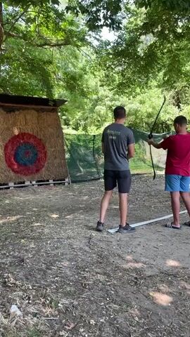 A video captures an archery practice session. In the foreground, two individuals stand facing a target made of hay bales with a concentric circle design. The person on the right is holding a bow and arrow, poised to shoot. To the left, another individual stands observing. In the background, a group of people are seated on benches, watching the activity. The environment appears to be an outdoor setting with trees and foliage providing shade. The ground is covered with dirt and fallen leaves.