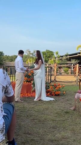 The video shows a wedding ceremony taking place outdoors. A man and a pregnant woman, presumably the bride and groom, stand facing each other holding hands. The man is wearing a white collared shirt and khaki pants with brown shoes. The woman is wearing a white dress with a side slit, a veil, and high heels. Behind them is a small table covered in an orange cloth, decorated with orange flowers and oranges. The background includes a metal and wood fence, trees, and what appears to be a stable with a horse visible inside. Several people are seated watching the ceremony. One person is visible to the left wearing a patterned shirt and jeans. Another person is partially visible to the right. The sky is clear and blue. The scene appears to be taking place on a grassy lawn.