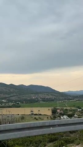 The video shows a view from a vehicle traveling along a road. In the foreground, there is a metal guardrail and some greenery. Beyond the guardrail, there is a rural landscape with fields, trees, a village, and rolling hills in the background. Power lines are visible stretching across the frame. The sky is cloudy, suggesting it is either early morning or late evening. The camera appears to be mounted on a car or bus, capturing the passing scenery. There are no people or animals clearly visible in the frame. The overall impression is a serene, pastoral setting observed from a moving vehicle.