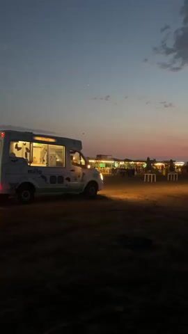 A white ice cream truck with a cartoon cow design and the text "milla" on its side is parked on a dirt lot at dusk. The truck has its lights on. In the background, there are several tables and chairs, some with umbrellas, and structures that appear to be small shops or food stalls. The sky is a gradient of deep blue at the top to pink and orange near the horizon, with some scattered clouds. The overall scene suggests a beachside or outdoor market setting as evening approaches.