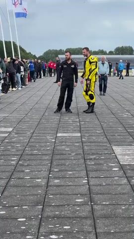 Two men stand facing each other on a paved area. The man on the left wears a black racing suit with a white logo on the chest. The man on the right wears a bright yellow and black racing suit with the number 6 visible on the back and the name DAM on the leg. A crowd of people stands in the background, lined up, and further back there are tents and a stadium structure. Two flags, one white, blue, and red, and another white, red, and blue, are visible on flagpoles in the background. The ground is made of large, grey, rectangular pavers. The sky is overcast.