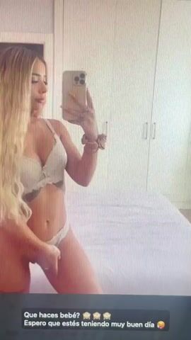 This is a video of a young woman taking a selfie in what appears to be a bedroom. She is wearing a white bra and underwear. The woman has long blonde hair and is posing for the camera. The room is brightly lit, and a bed is visible in the background. A phone is being held up to take the picture. The video is short and loops. At the bottom of the screen, there is text that reads "Que haces bebé? Espero que estés teniendo muy buen día," which translates to "What are you doing baby? I hope you are having a very good day." The context provided for this media file is "Mantov, Czechia," however, there are no visual cues in the video to confirm this location. The time of day is not specified but appears to be daytime due to the bright lighting. The overall emotion conveyed is playful and flirty.