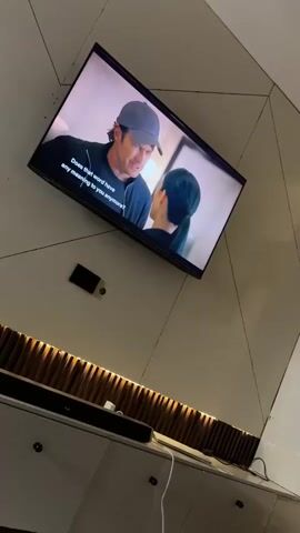 The video shows a television displaying a scene with a man wearing a baseball cap and a woman. The man is asking, "Does that word have any meaning to you anymore?". The text on the screen then changes to "How have you gotten so good at lying". The television is mounted on a wall with a wooden design below. A remote control and a coin are lying on the floor near the wall.