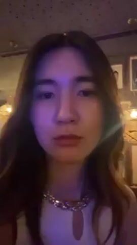 A young woman is shown in a close-up selfie video. She has long, straight, dark hair and fair skin. She is wearing a white top with a silver, chunky necklace. She winks and makes a kissing face at the camera. The background is a dimly lit bar or restaurant, with framed pictures on the wall behind her. The lighting in the video appears to be purple or blue-toned. Music is playing in the background.