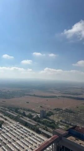 This is a vertical video shot from a high vantage point, likely a building or structure, looking out over a vast industrial landscape under a bright blue sky with scattered white clouds. The camera pans upward to reveal a tall, concrete industrial chimney on the right side of the frame. The chimney is partially obscured by the camera's perspective, but its sheer scale is evident.

In the foreground, to the left of the chimney, a railing of a structure is visible, suggesting the vantage point is from another elevated platform. Below, the industrial complex stretches out. Rows of what appear to be white storage containers or railway cars are neatly arranged in the lower left section of the image. Adjacent to these are buildings with reddish-brown roofs and industrial infrastructure, including a network of power lines and electrical towers.

The middle ground features a sprawling, somewhat desolate landscape. Patches of brown and green farmland are interspersed with what looks like abandoned or undeveloped land. A few scattered buildings, including a multi-story structure, are visible within this area. The background fades into a hazy, distant horizon, suggesting a large, open area with rolling hills or mountains in the far distance, softened by atmospheric conditions. The overall impression is one of an industrial zone situated within a rural or semi-rural environment, under a clear, sunny day.