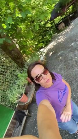 A woman is filming herself in a wooded area, holding a phone in her right hand and extending her arm to capture a selfie-style video. She is wearing a purple t-shirt with blue lettering that reads "Queen," blue jeans, and sunglasses. The woman is looking at the camera and appears to be talking or singing. She is standing on a gravel path, with trees and greenery surrounding her. To her left, there is a wooden bench with several potted plants. The video is shot from a low angle, looking up at the woman.