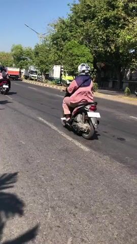 The video shows a street scene with several vehicles and trees. In the foreground, a portion of a motorcycle's mirror is visible, reflecting another motorcycle. Further down the road, a person wearing a headscarf and a pink top is riding a motorcycle. To the right of this motorcycle, a green and white truck is parked on the side of the road. In the background, more trees and buildings can be seen. The weather appears to be sunny.
