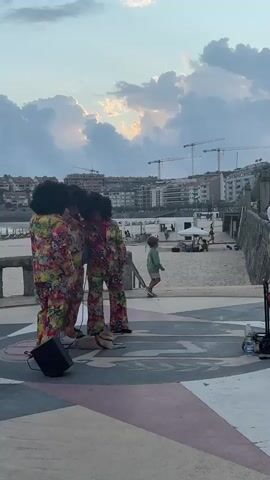 A group of three performers in colorful, patterned suits with large afro wigs are standing in a plaza near a beach. One performer is holding a microphone and appears to be singing or speaking into it. A small black speaker is placed on the ground in front of them. In the background, a beach can be seen with several people walking and relaxing. Buildings line the street behind the beach, and construction cranes are visible in the distance. A young boy in a green shirt and shorts is running across the plaza, and a young girl in a green shirt and white shorts is standing nearby, watching the performers. A man in a white shirt and hat is also present, facing the performers and gesturing with his hands. The sky is cloudy with patches of sunlight breaking through.