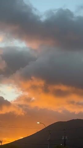 The video shows a sky with dramatic clouds at sunset. The clouds are a mix of dark gray and vibrant orange, indicating the sun is low on the horizon and casting a warm glow. A silhouette of a mountain range is visible in the background. In the foreground, there are dark silhouettes of trees and some utility poles with wires. The overall impression is of a natural landscape during twilight.
