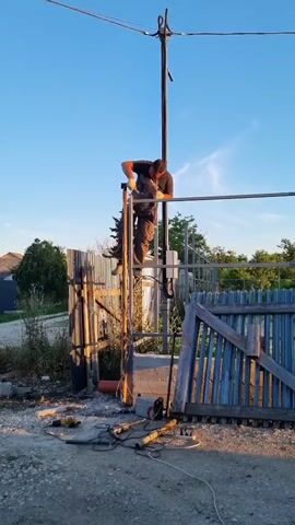 A man is working on a metal structure in an outdoor setting. He is standing on a scaffold-like platform, holding what appears to be a metal rod. He is wearing a dark t-shirt and pants. Several power tools, including grinders and drills, are laid out on the ground in front of a fence. The scene is illuminated by what seems to be late afternoon or early evening sun, casting long shadows. The sky is a clear, bright blue. Behind the man is a tall pole with wires running across it, possibly a utility pole. The setting appears to be a rural or semi-rural area, with houses and trees visible in the background. The overall impression is one of construction or repair work in progress.