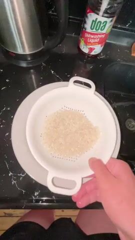 A person's hands are visible as they hold a white colander filled with white rice. The colander is over a white plate with a floral design. The person then moves the colander over a sink. A bottle of dish soap is on the counter behind the colander. A metal kettle is also visible behind the colander. The sink is dark and has a drain.