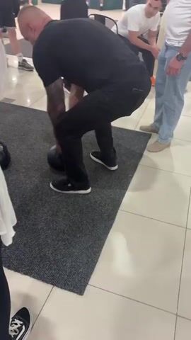 A man in a black t-shirt and black pants is holding a large black kettlebell in a shopping mall. He is standing on a dark gray mat. To his left, a person wearing a white garment is partially visible. To his right, another man in a white t-shirt and blue jeans is standing. In the background, other shoppers and store displays are visible. The man with the kettlebell is the central focus of the video. He lifts the kettlebell up to his chest and then extends his arms forward.